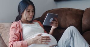 A pregnant woman is relaxing on a brown couch, gently resting her hand on her belly while looking at a tablet in her other hand. She appears calm and focused, enjoying a quiet moment at home. A pregnant woman is relaxing on a brown couch, gently resting her hand on her belly while looking at a tablet in her other hand. She appears calm and focused, enjoying a quiet moment at home.